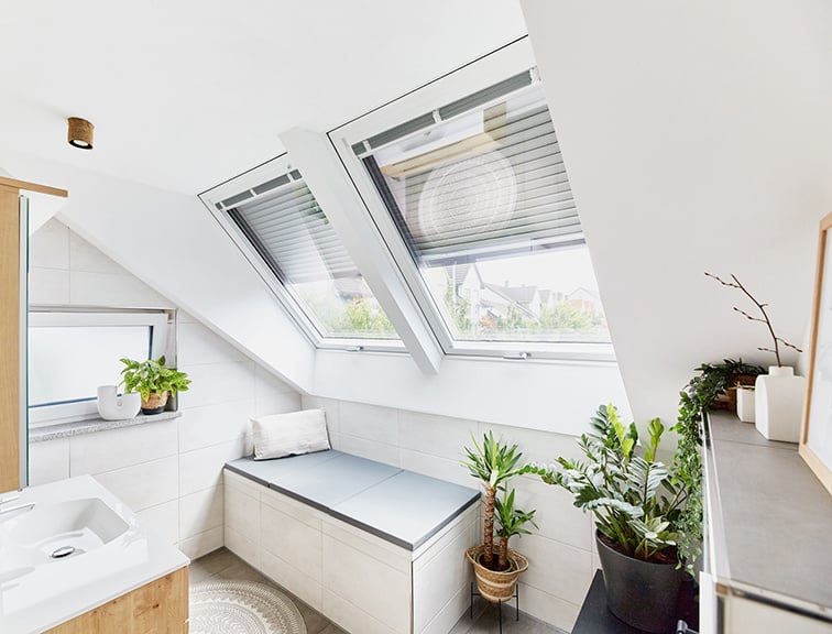 Badezimmer mit zwei Roto Dachfenstern, einem Waschbecken mit Spiegelschrank und einer Sitzmöglichkeit unter dem Dachfenster.