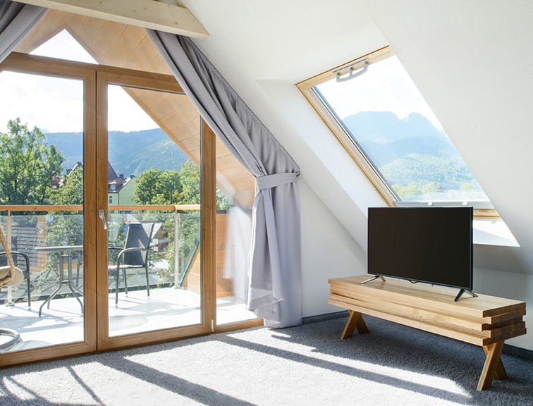 Dachgeschosszimmer mit Blick auf einen Balkon mit großer Fensterfront und einem Roto Dachfenster mit einem Lowboard und Fernseher darunter.