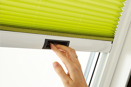 Hand zieht ein hellgrünes Faltstore nach unten, welches an einem Roto Dachfenster innen angebracht ist